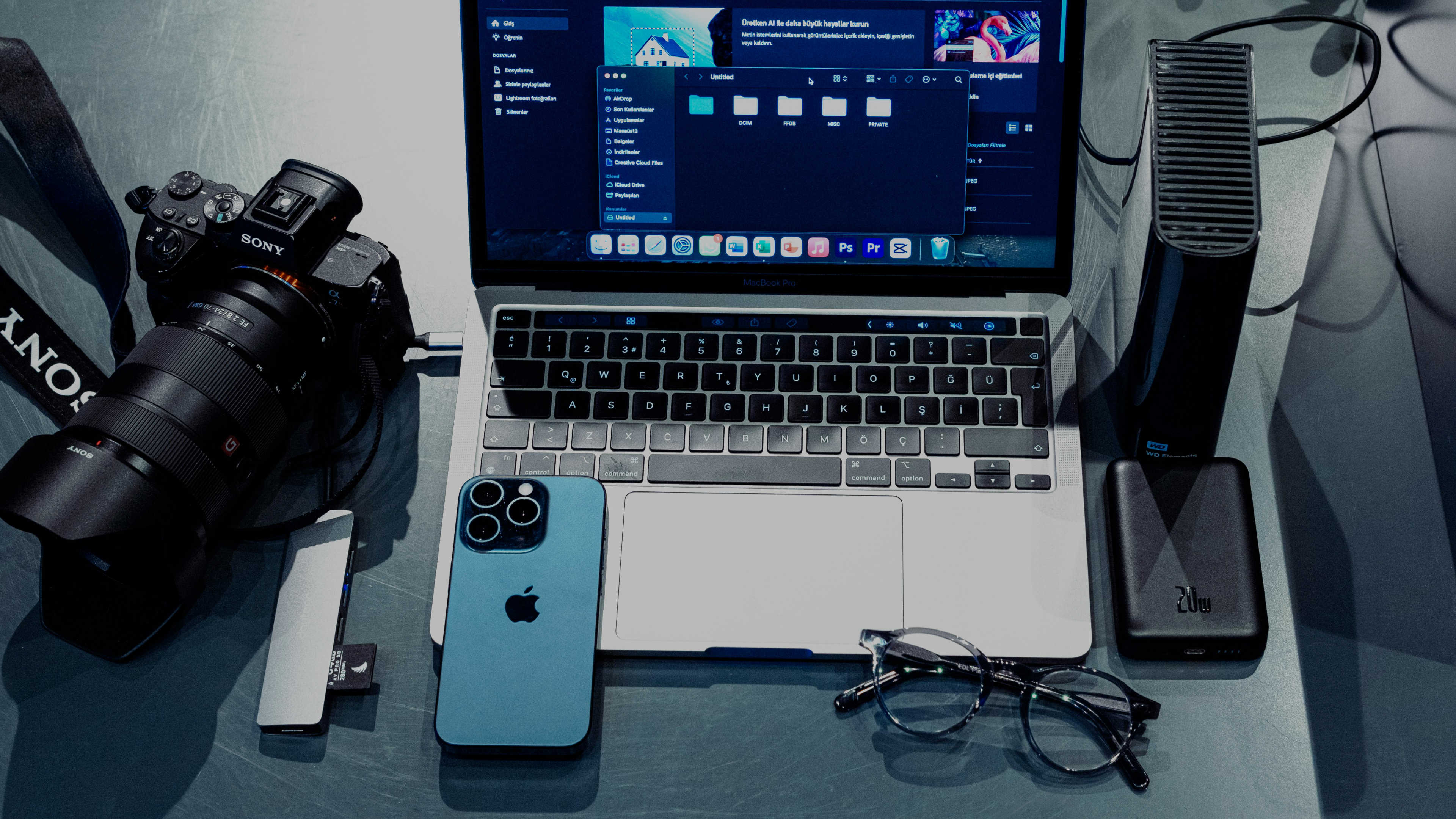 A desk with a camera, phone, laptop, glasses and external drives neatly arranged for organizing travel files