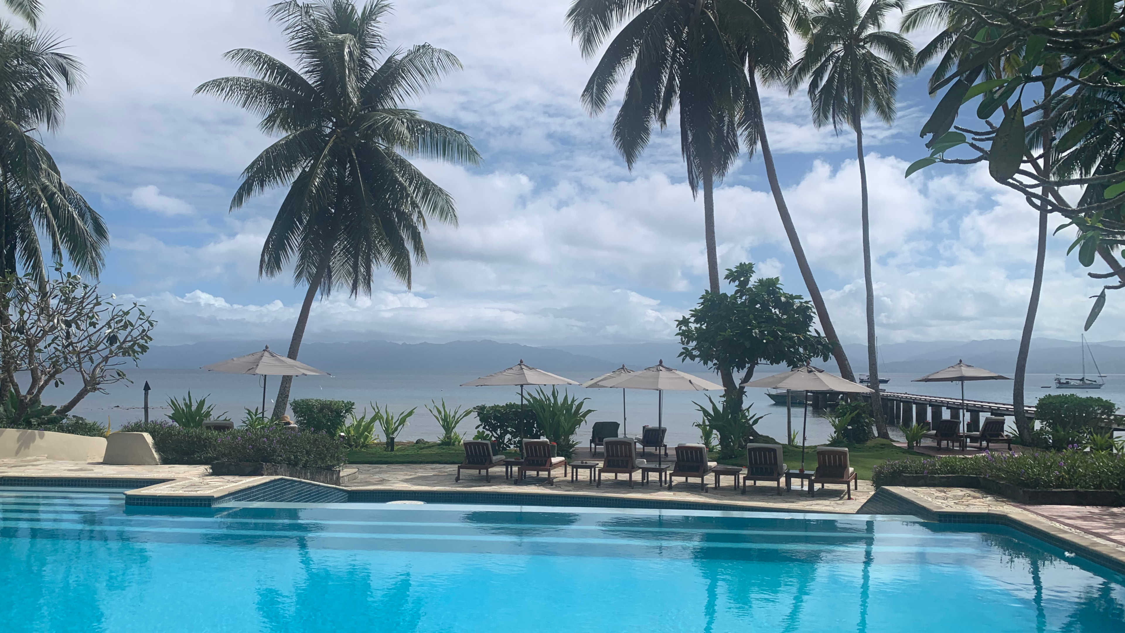 An infinity pool with chaise lounges and beds surrounding it, with a gorgeous seascape behind of Savusavu Bay