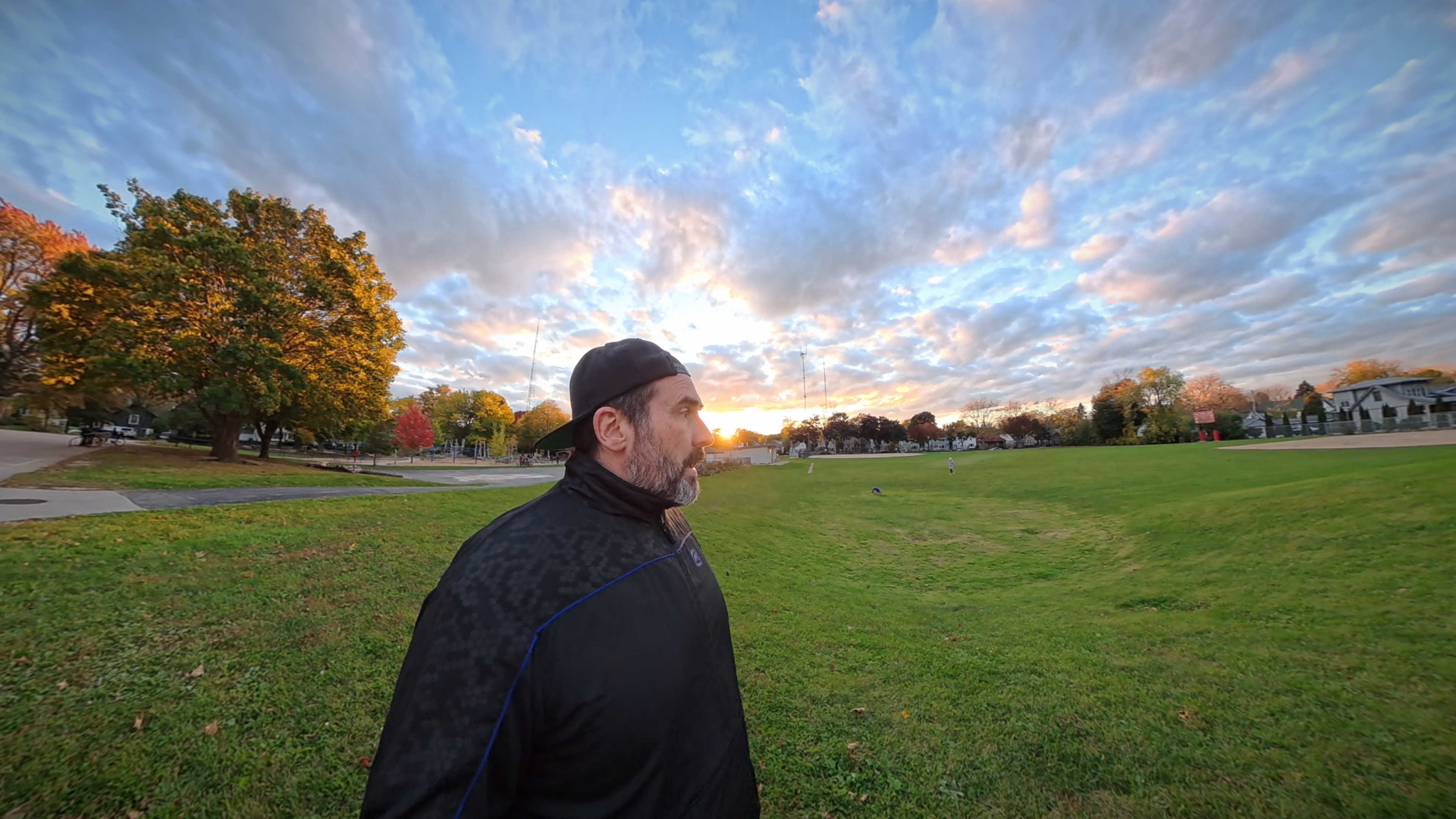 Me wearing a backwards baseball cap with a grassy playground, the sunset and evening sky in the background